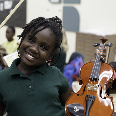 A girl in a green polo shirt smiles while she is holding up a violin